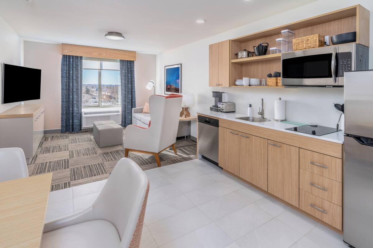 A modern hotel room with a dining area, kitchen, and a large window with a city view.