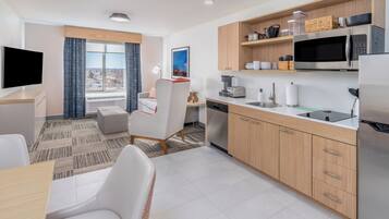 A modern hotel room with a dining area, kitchen, and a large window with a city view.