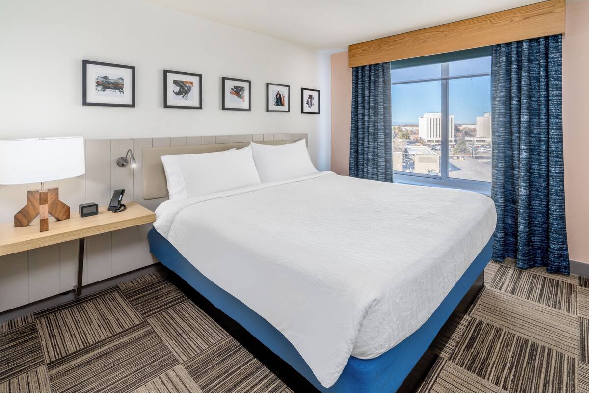 A hotel room with a large bed, a desk with a lamp, a phone, and framed pictures on the wall.