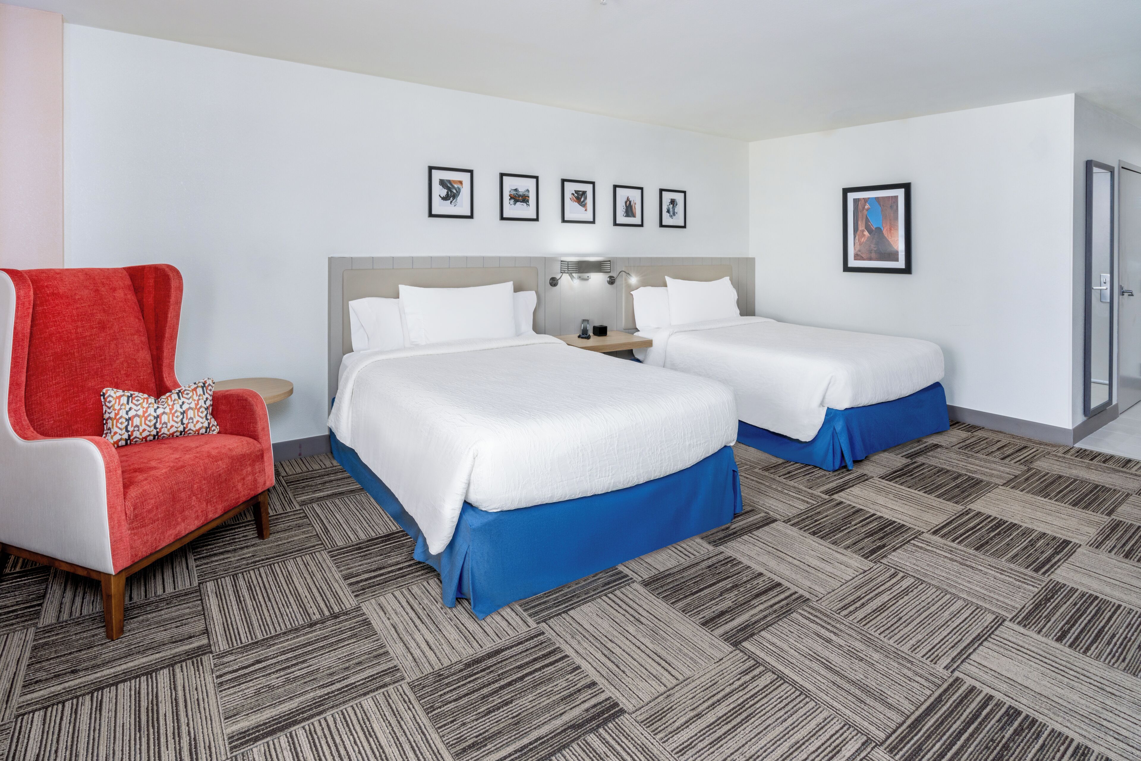 A hotel room with two beds, a red armchair, a nightstand with a lamp, and a framed picture on the wall.