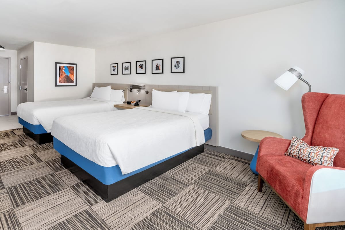 A hotel room with two beds, a red armchair, a small round table, and a wall-mounted lamp.