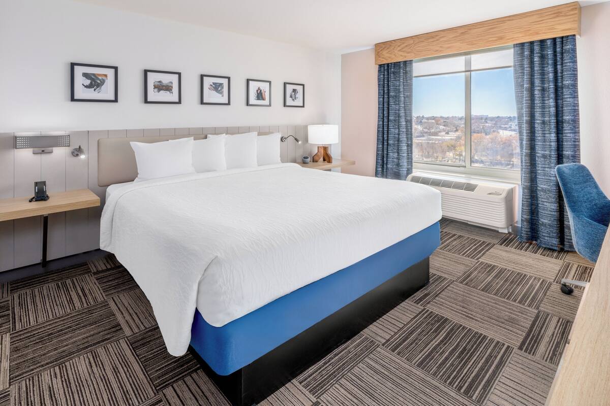 A hotel room with a large bed, a desk, a chair, and a window with a city view.