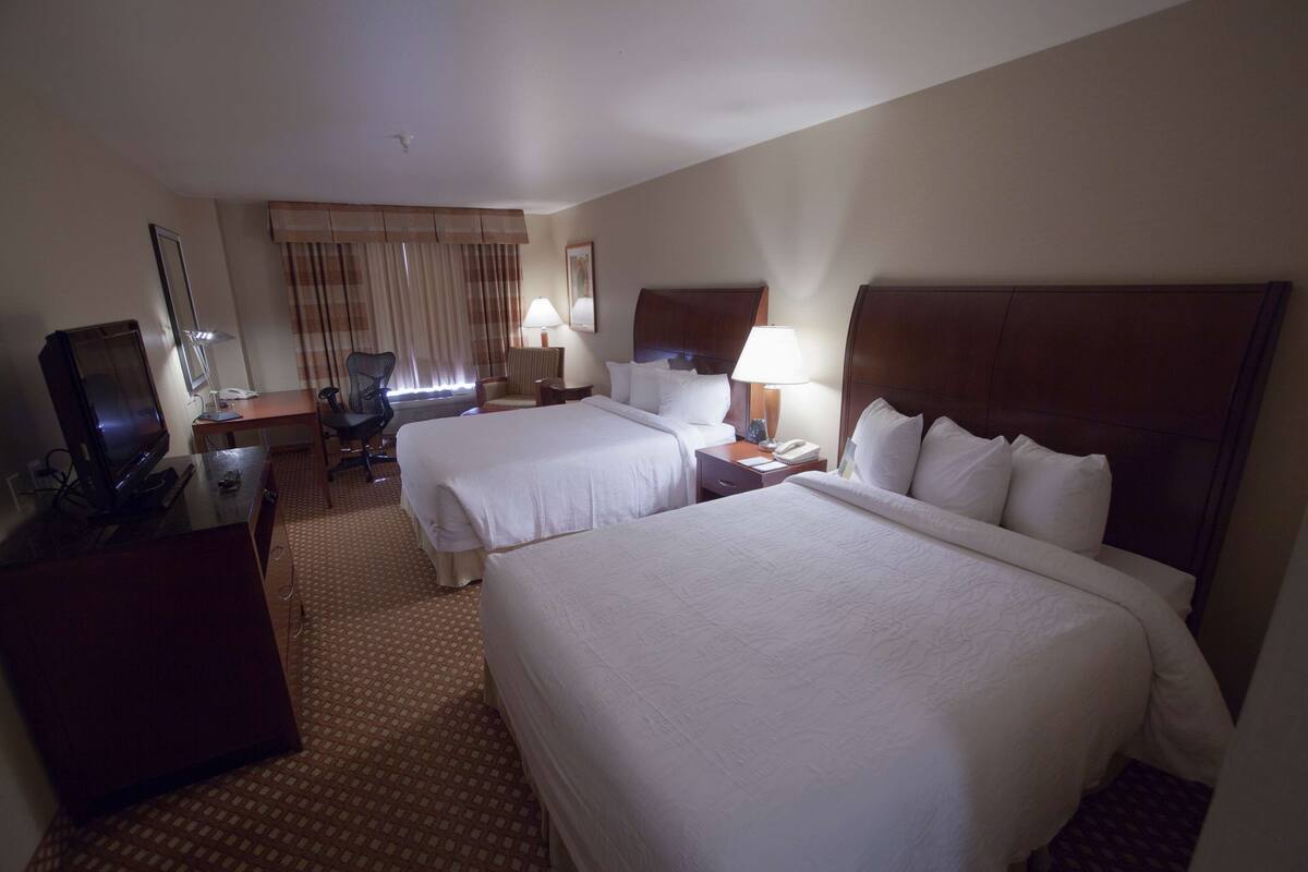 A hotel room with two beds, a desk, a chair, a TV, and a window with curtains.
