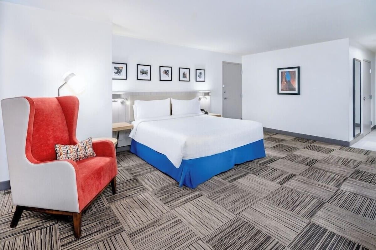 A hotel room with a bed, a red armchair, a nightstand, and a wall with framed pictures.