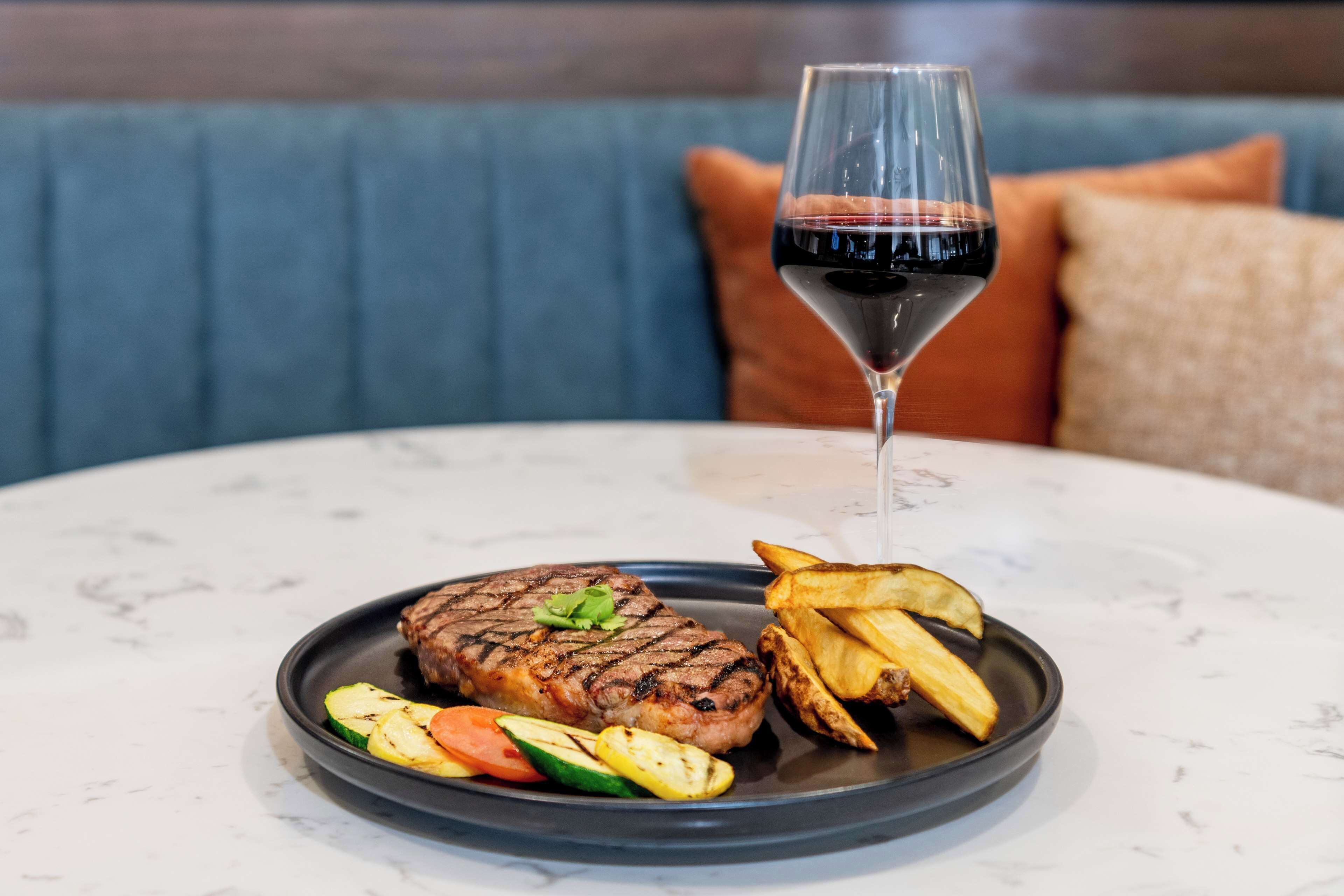 A steak with grilled vegetables and fries on a plate, accompanied by a glass of red wine, set on a round table with a blue couch in the background.