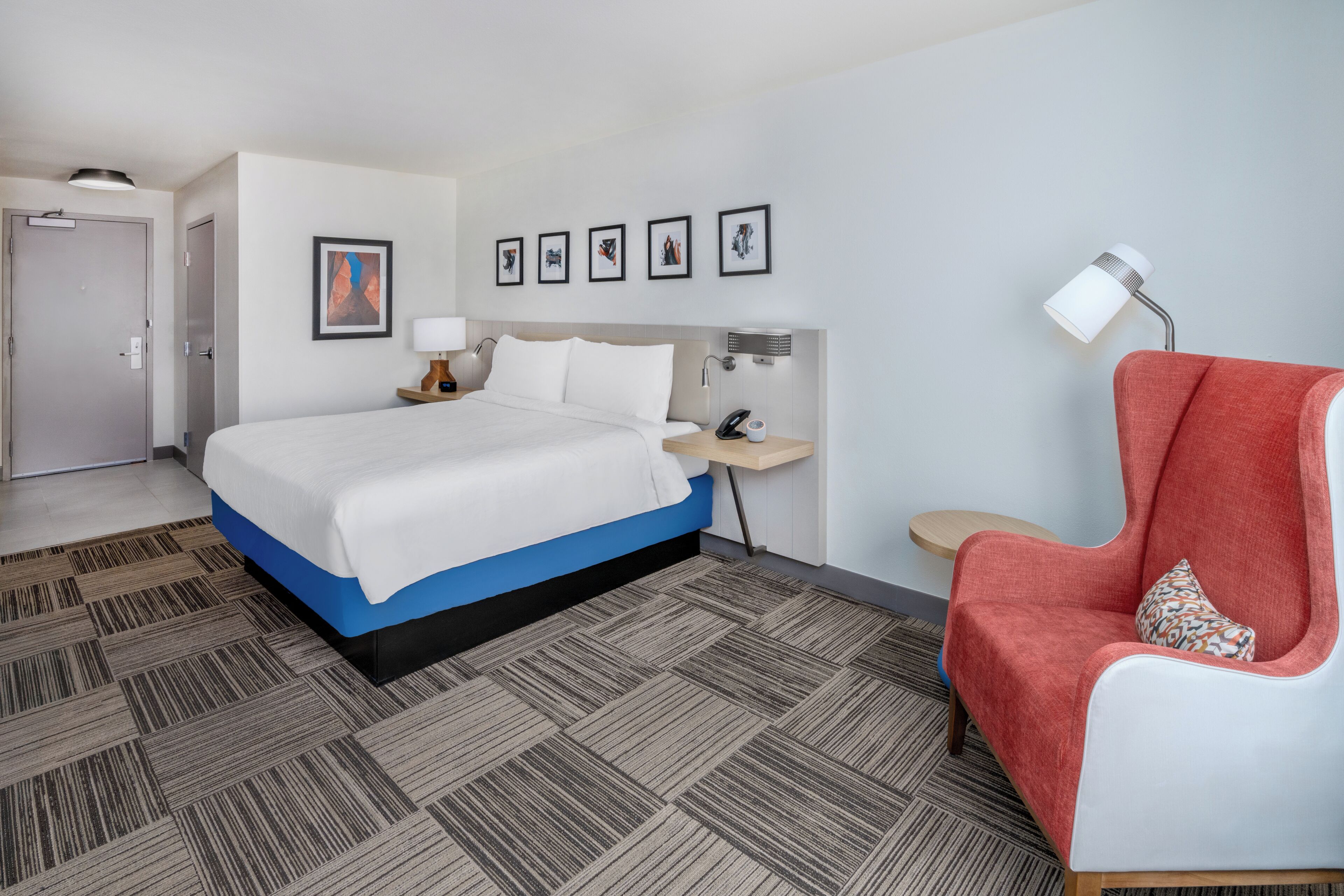 Une chambre d’hôtel avec un lit, un fauteuil rouge, une table de chevet avec une lampe, un téléviseur fixé au mur et des œuvres d’art encadrées.