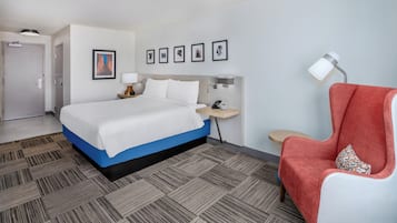 Une chambre d’hôtel avec un lit, un fauteuil rouge, une table de chevet avec une lampe, un téléviseur fixé au mur et des œuvres d’art encadrées.