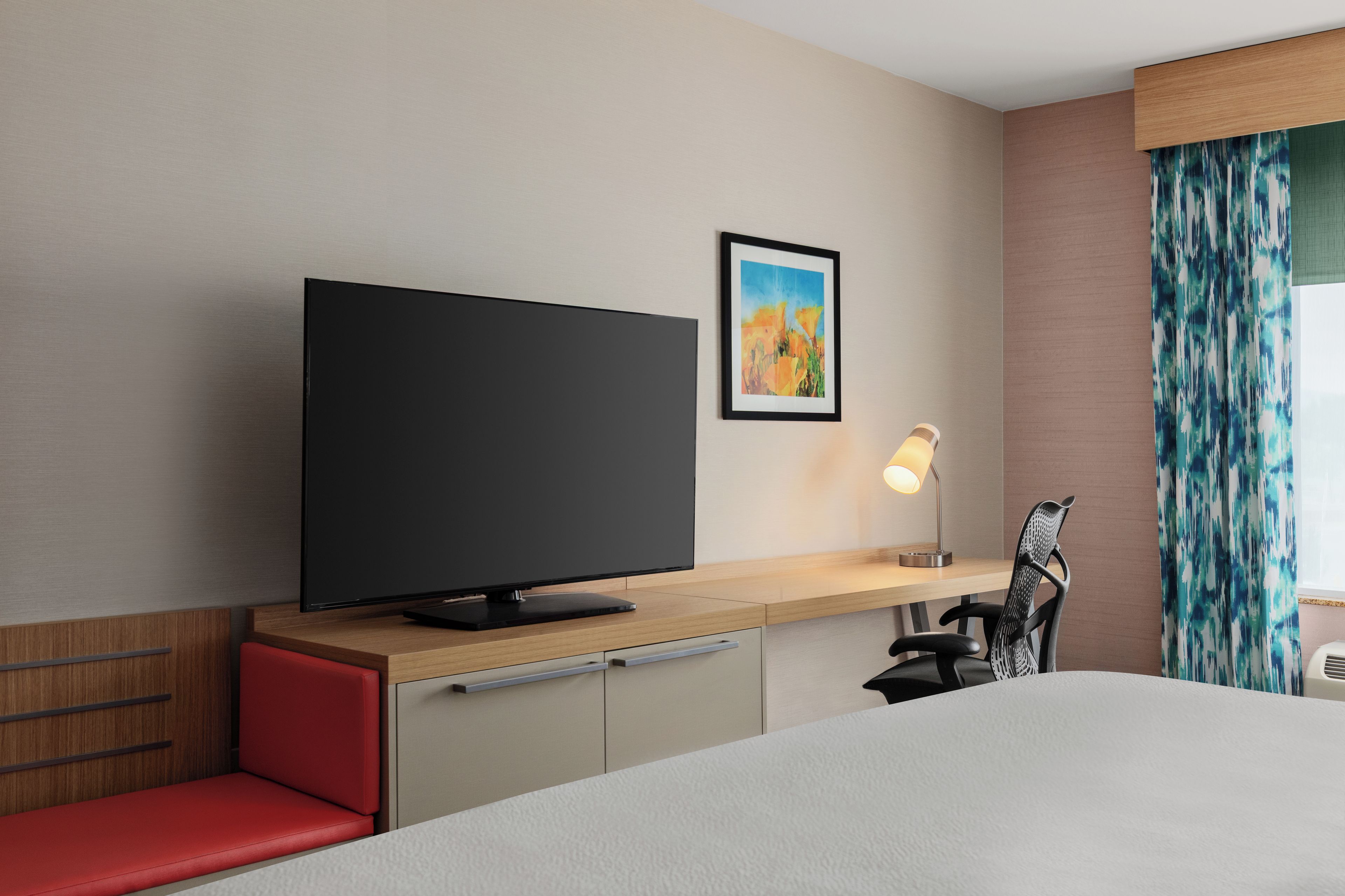 A hotel room with a flat-screen TV, a desk with a lamp, a chair, and a bed.