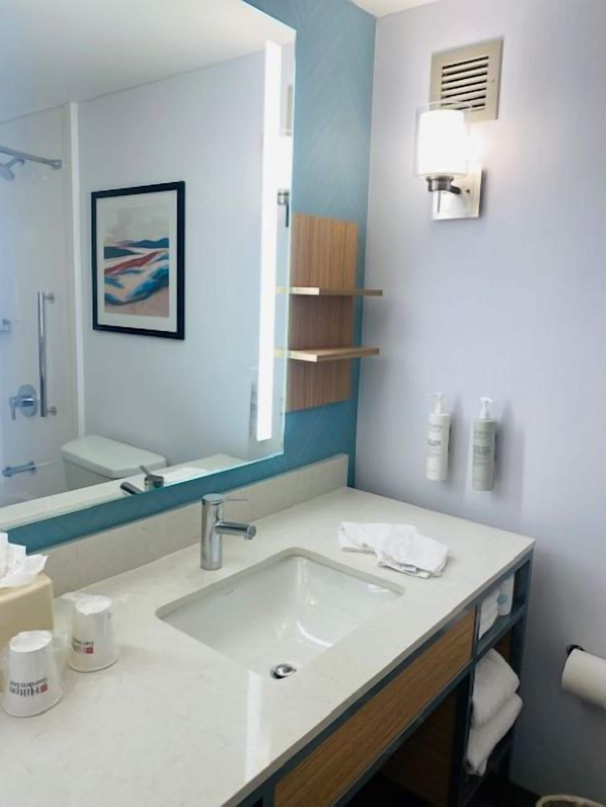 A modern bathroom with a large mirror, a sink, a towel rack, and a shelf with toiletries.
