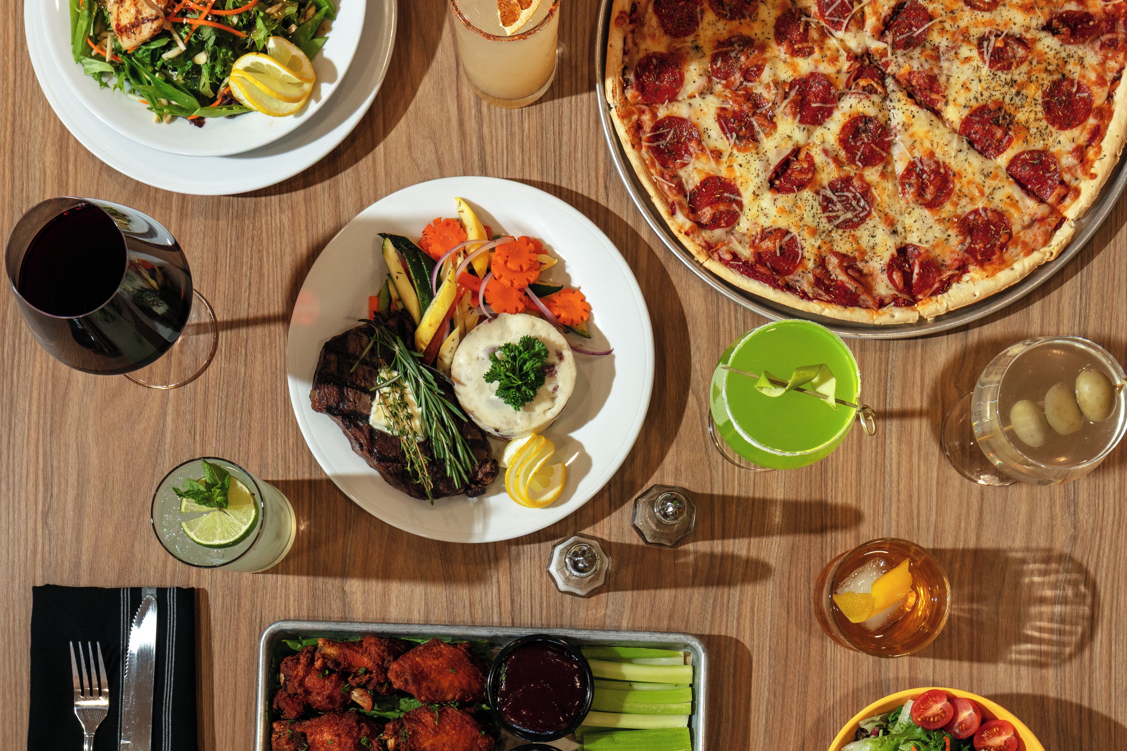 Una mesa de comedor con diversos platos, como pizza, ensalada y carne, además de bebidas y cubiertos.
