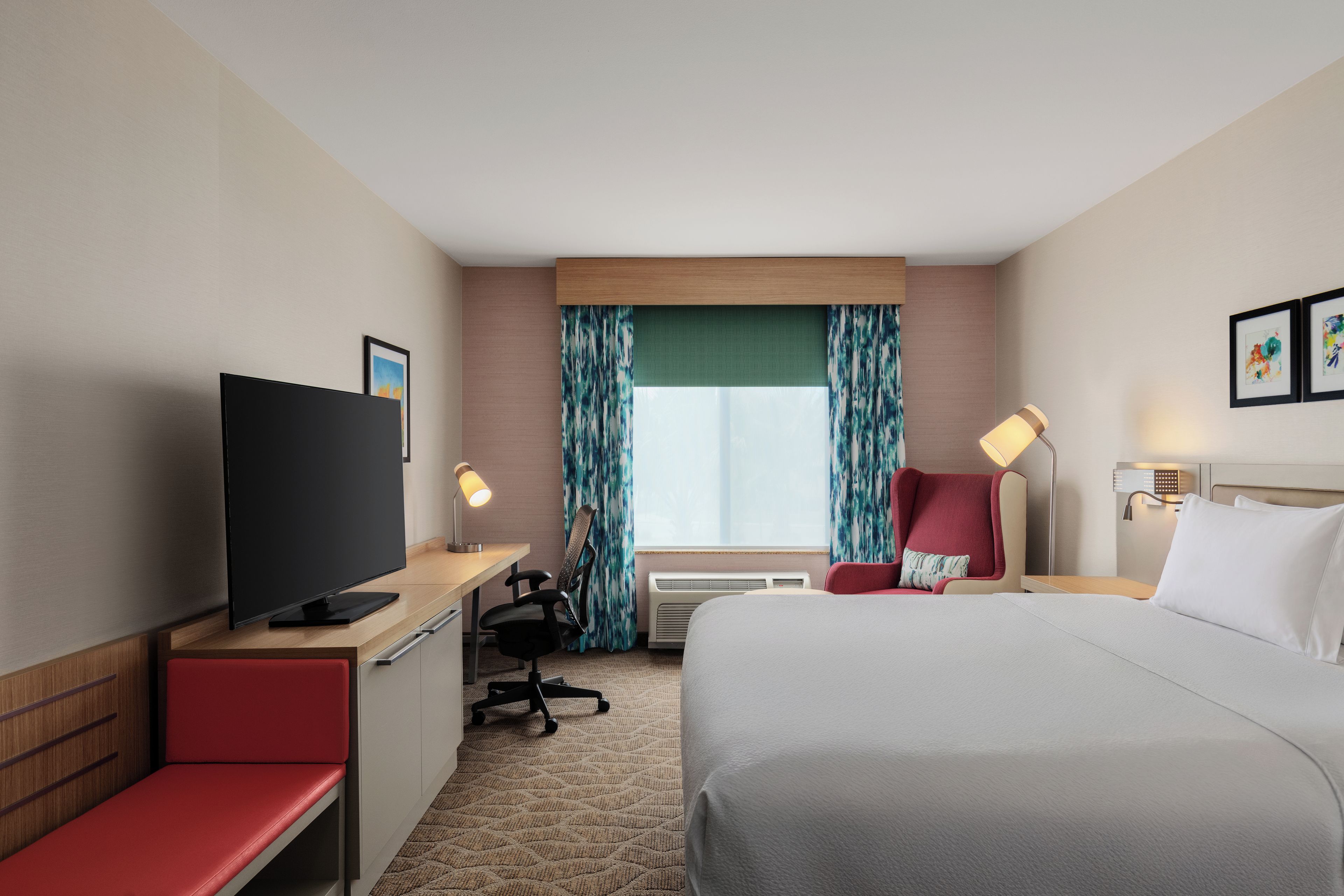 Une chambre d’hôtel comprenant un lit, un bureau avec une télévision, une chaise, une lampe de bureau et une fenêtre avec des rideaux.