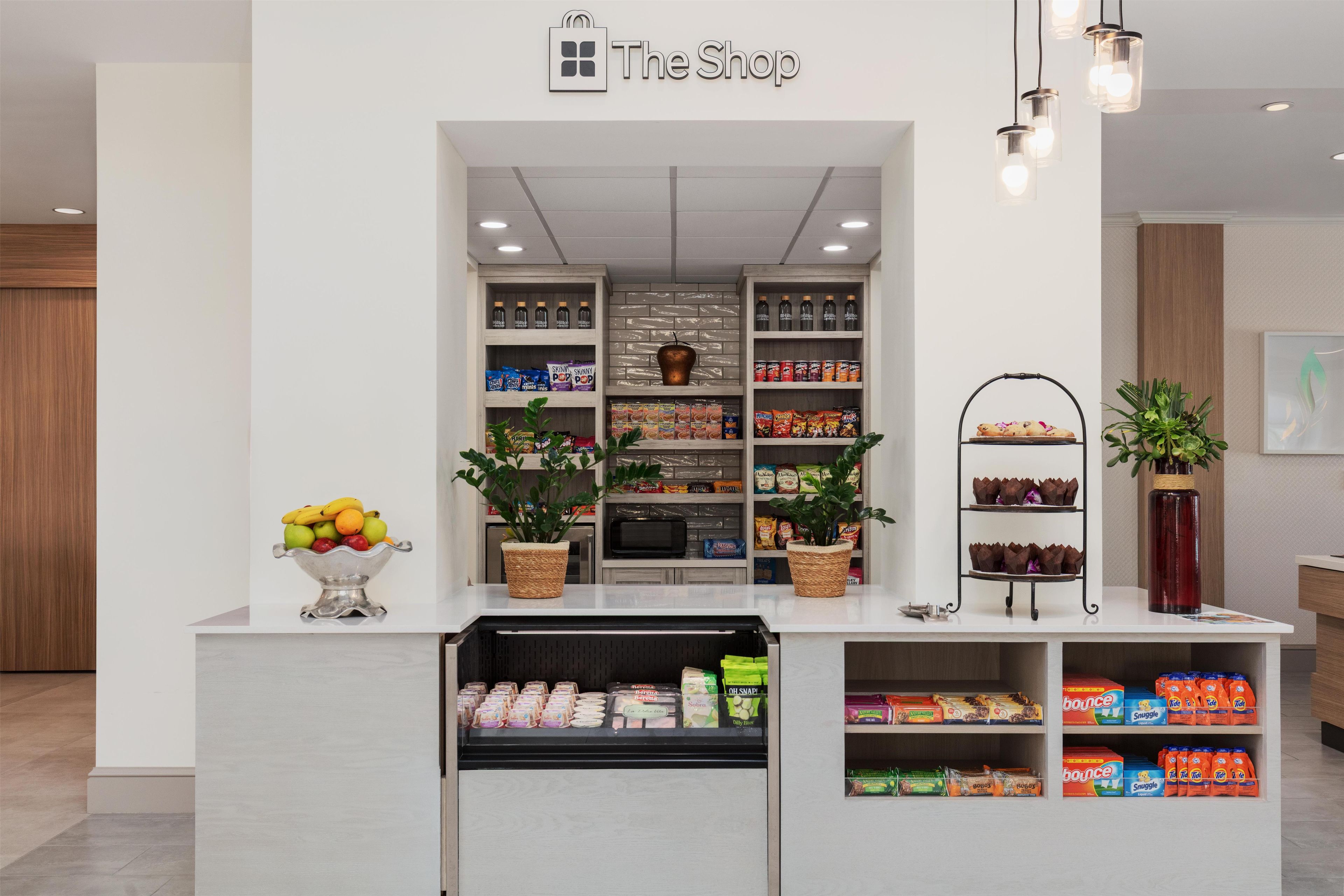 L’intérieur d’un magasin, avec un comptoir blanc, des étagères remplies d’en-cas, un panier de fruits et un présentoir de boissons en bouteille.