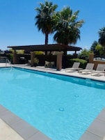 Una piscina rectangular con agua azul cristalina, rodeada de sillas de descanso y una zona de estar cubierta, con palmeras al fondo.
