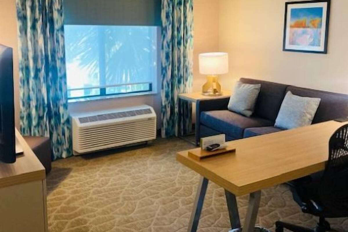 A hotel room with a bed, a desk, a chair, a lamp, a TV, a window with curtains, and a painting on the wall.