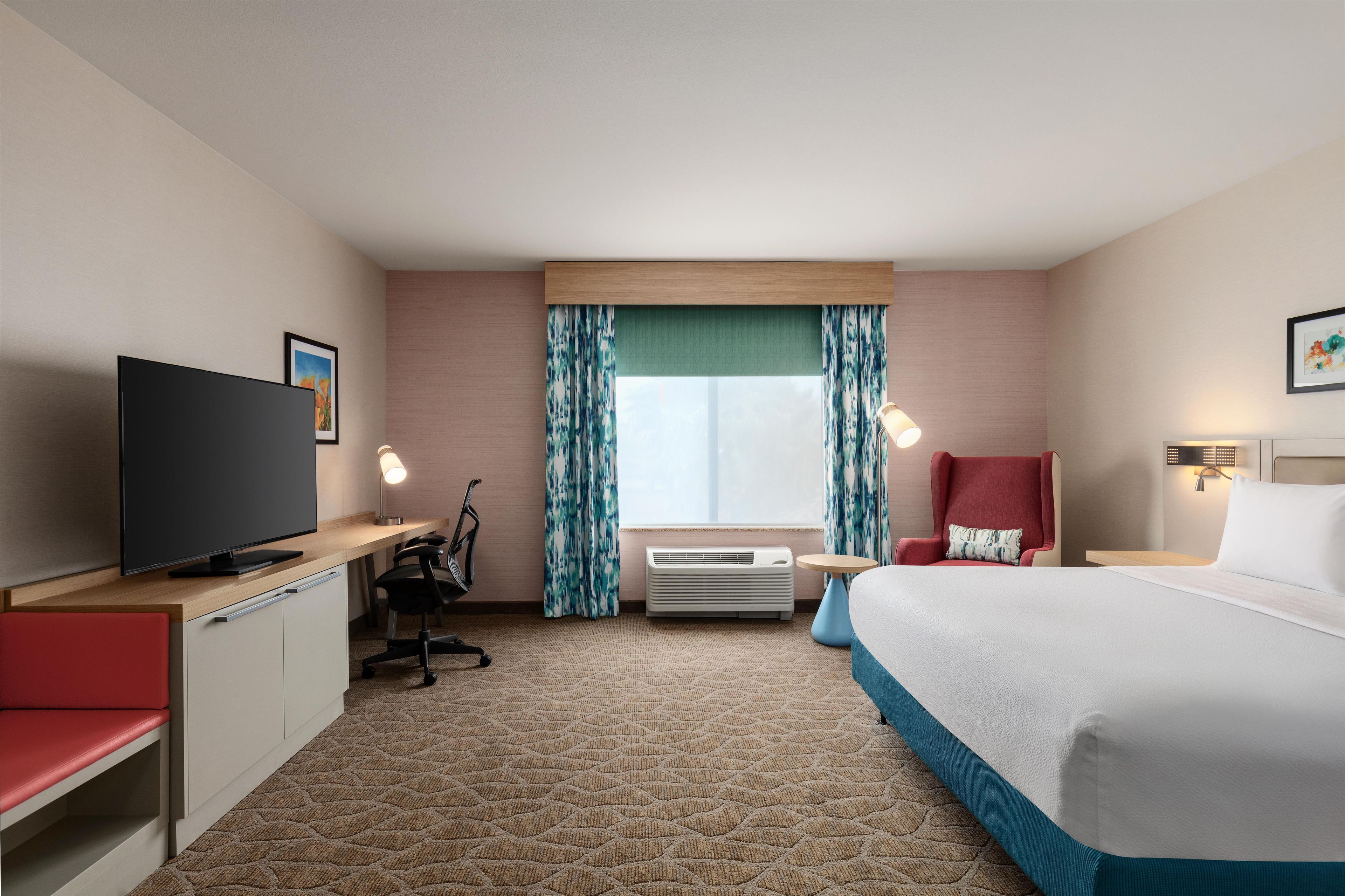 Habitación de hotel con cama, escritorio con televisor, una silla, lámpara de escritorio, ventana con cortinas y equipo de aire acondicionado.