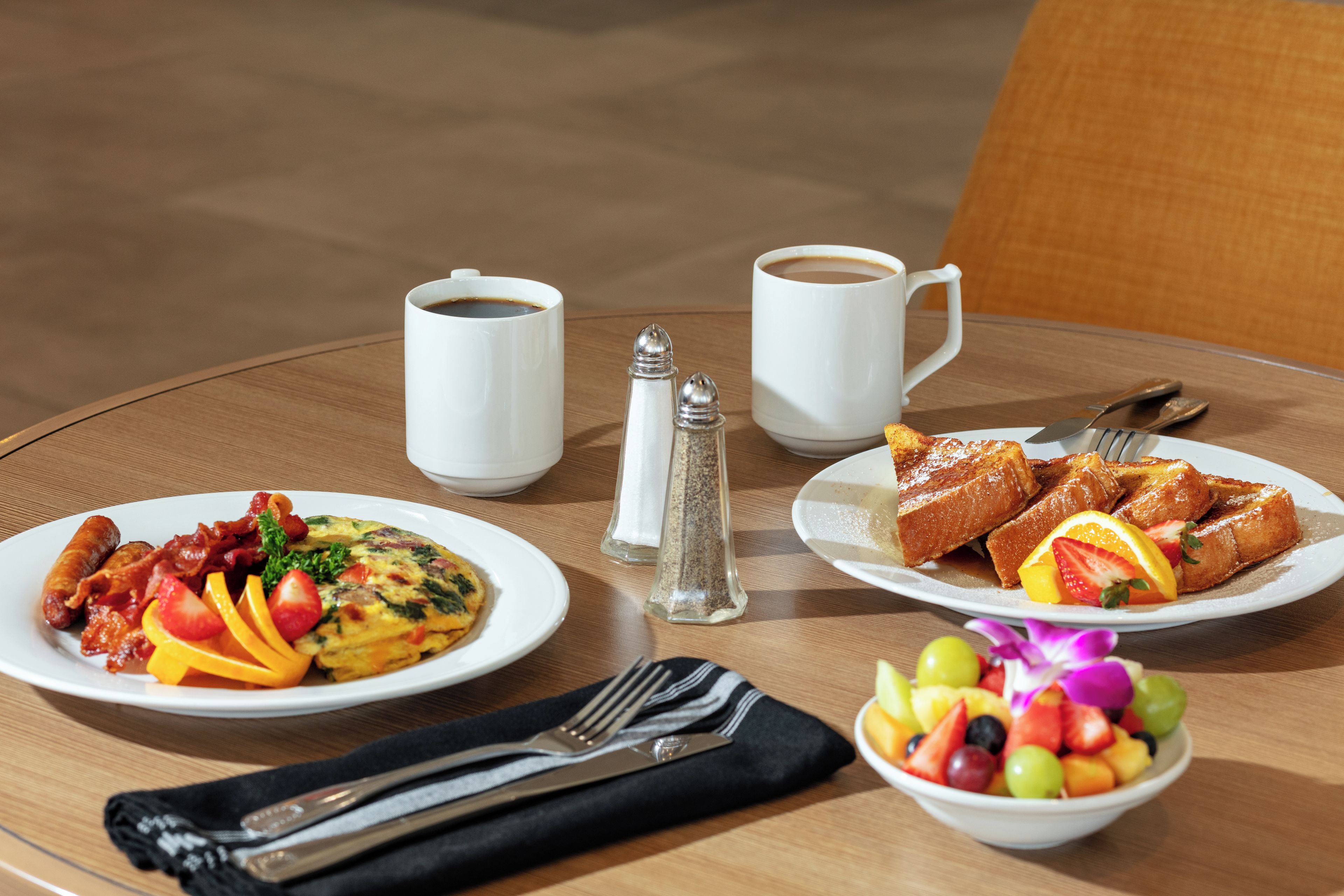 Una escena de desayuno con un plato de huevos, tocino y fruta, un bol de fruta mixta, dos tazas de café y un salero sobre una mesa redonda.