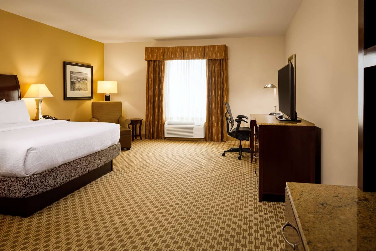 A hotel room with a large bed, a desk with a computer, a chair, a television, and a window with curtains.