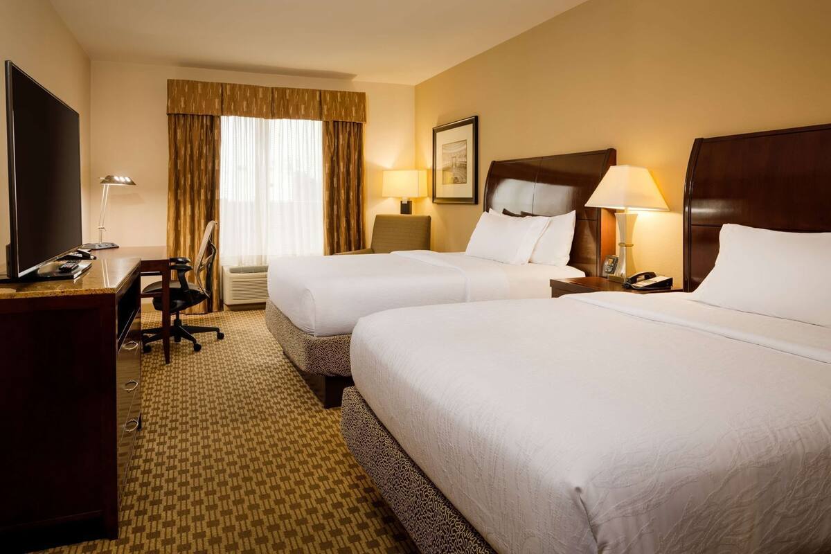 A hotel room with two beds, a desk, a chair, a TV, and a lamp.