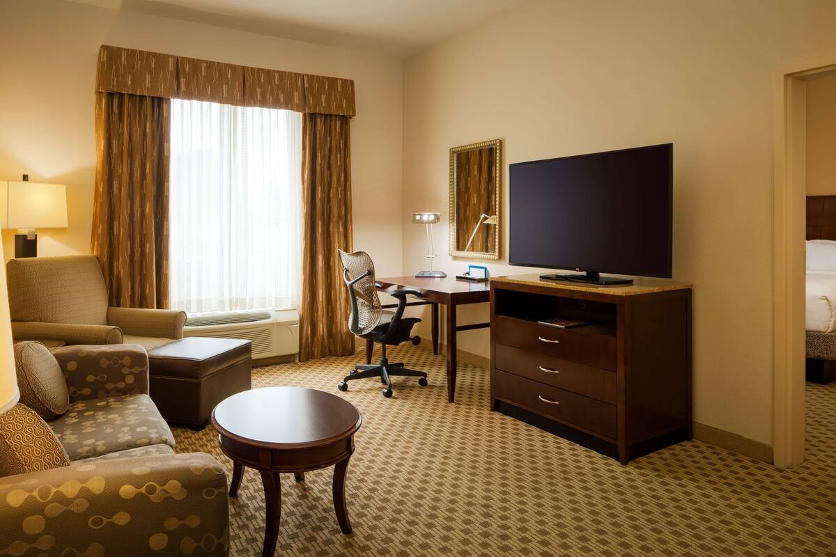 A hotel room with a sofa, a desk, a chair, a television, and a lamp.