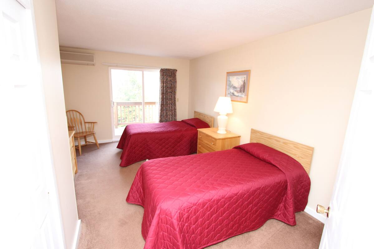 A room with two beds, a chair, a nightstand with a lamp, and a window with curtains.