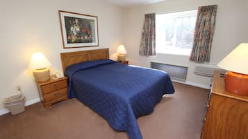 Een slaapkamer met een bed, nachtkastjes, een lamp, een prullenbak, een radiator en een raam met gordijnen.