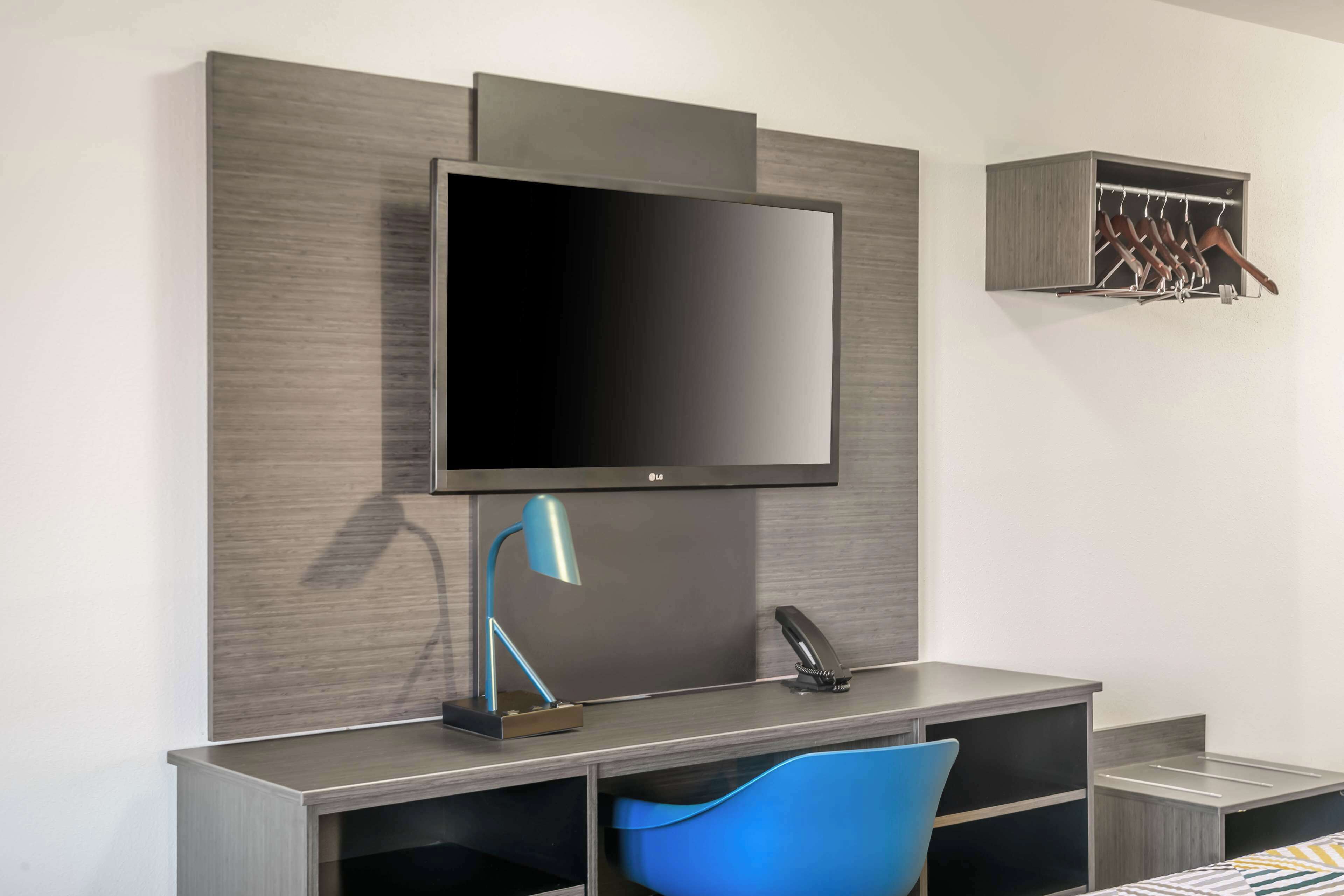 A modern room with a flat-screen TV mounted on a wall, a desk with a lamp, a chair, and a coat rack.