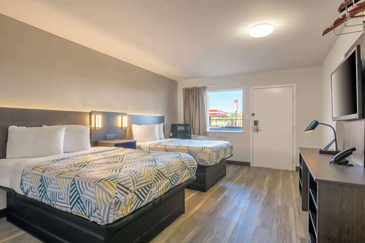 A hotel room with two beds, a desk, a TV, and a window with a view of a building.