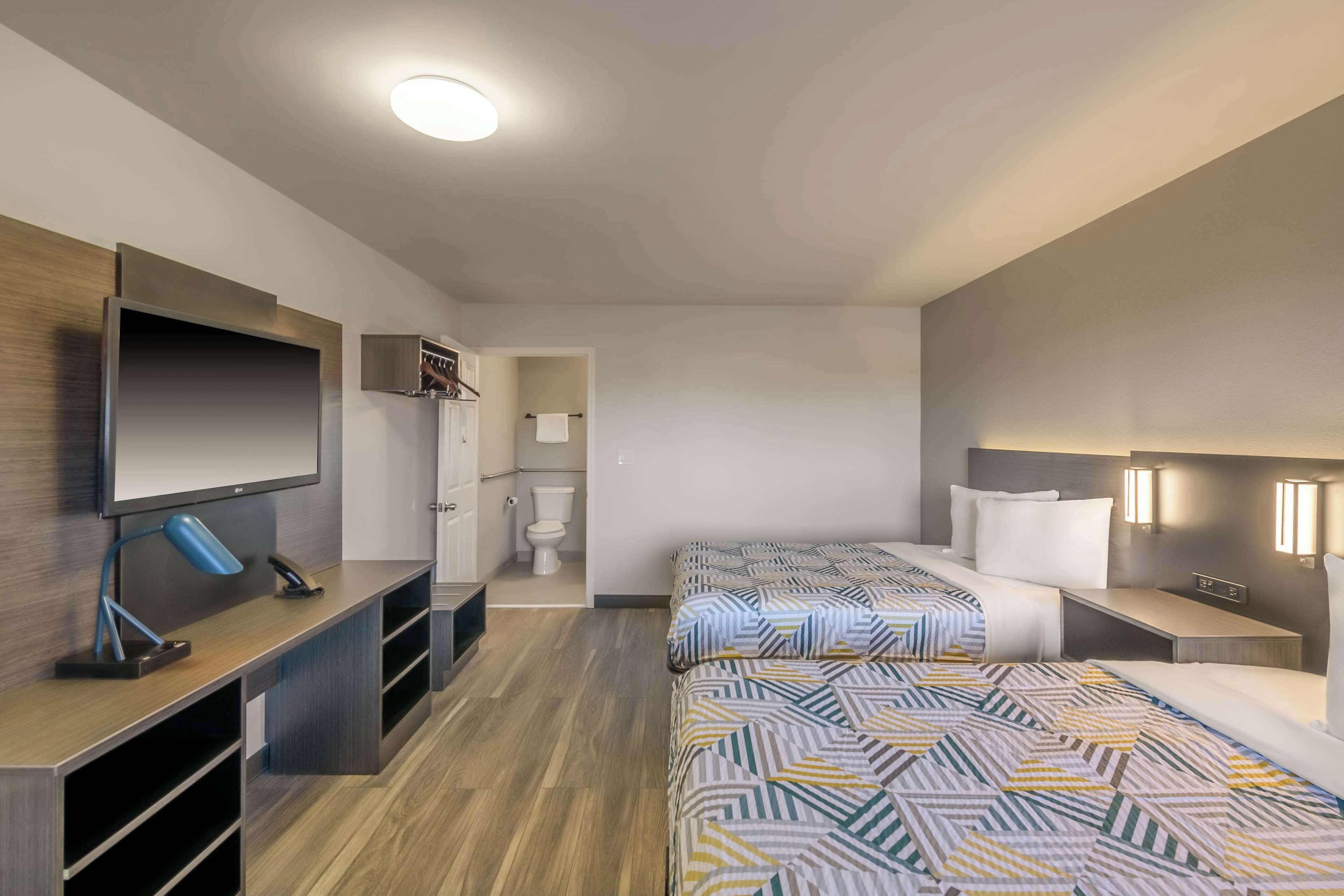 A modern hotel room with a flat-screen TV, a desk with a lamp, two beds with patterned bedding, and a small bathroom.