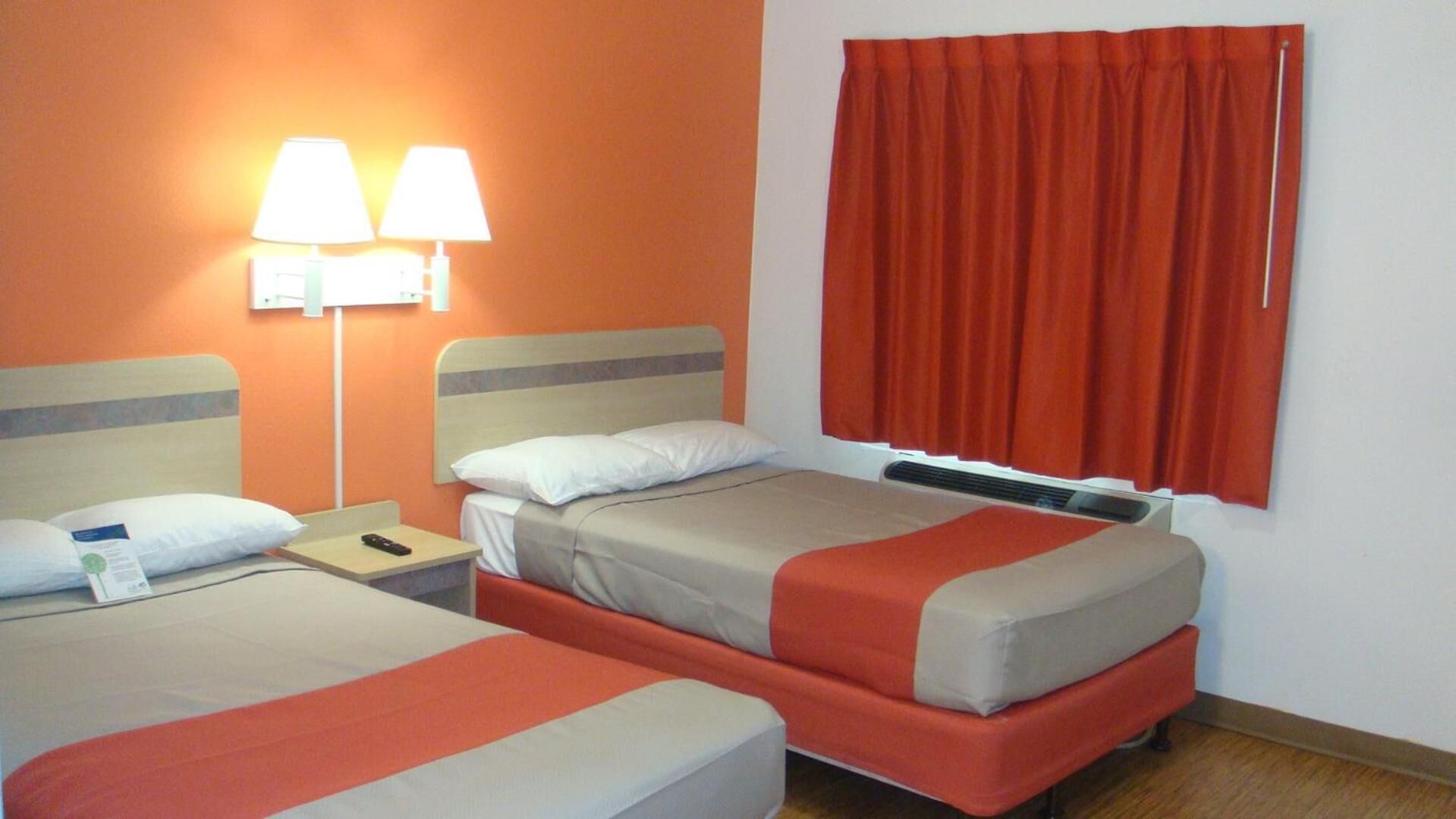 A hotel room with two beds, a nightstand, a lamp, and a window with curtains.