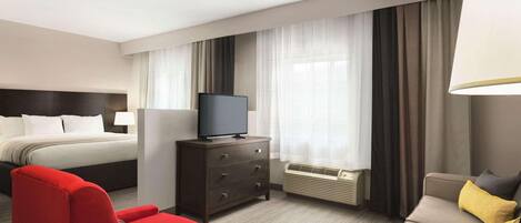 Habitación de hotel con cama, televisor en una cómoda, una silla roja, sofá color beige y una mesita.