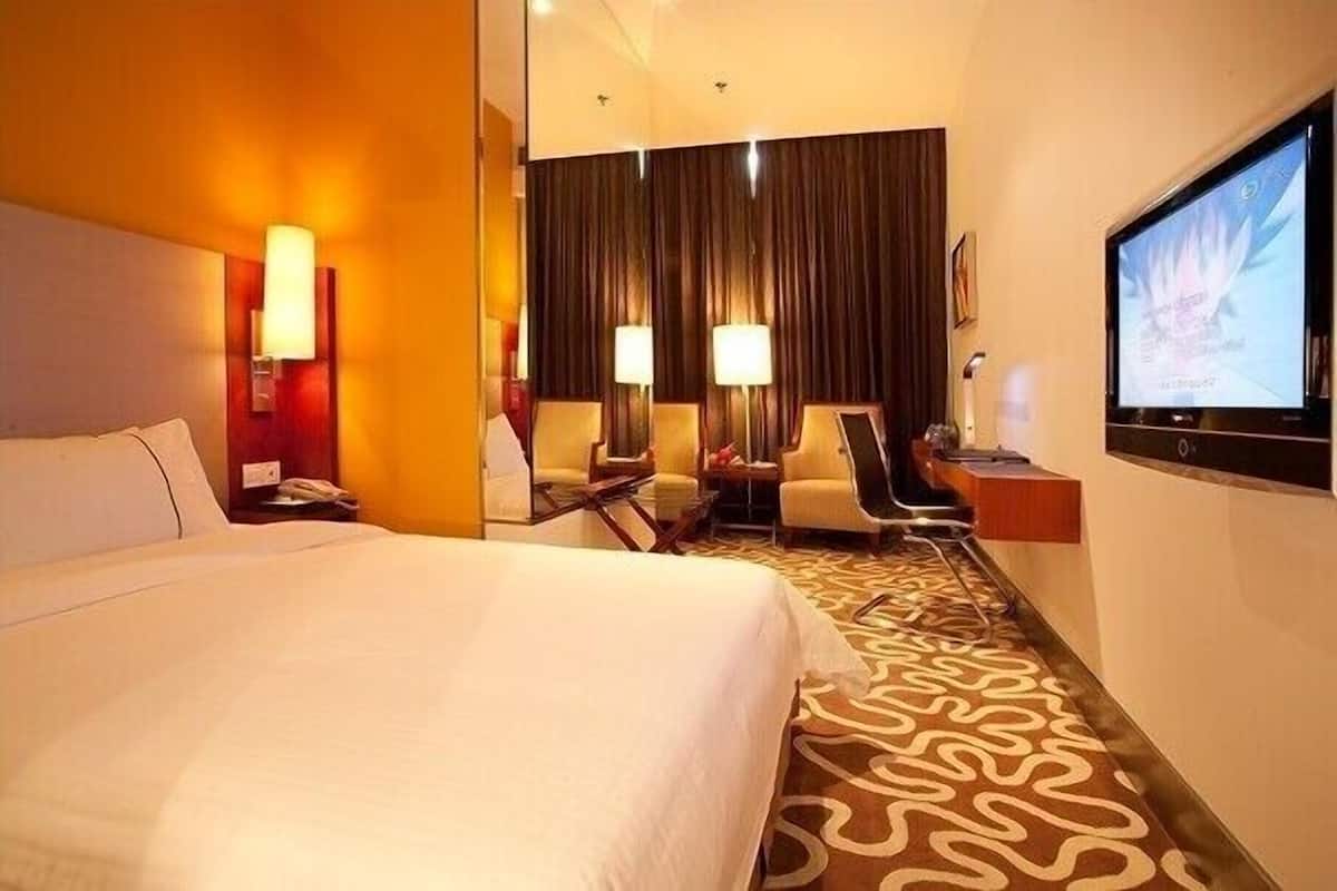 A hotel room with a large bed, two armchairs, a desk with a lamp, a television, and a patterned carpet.