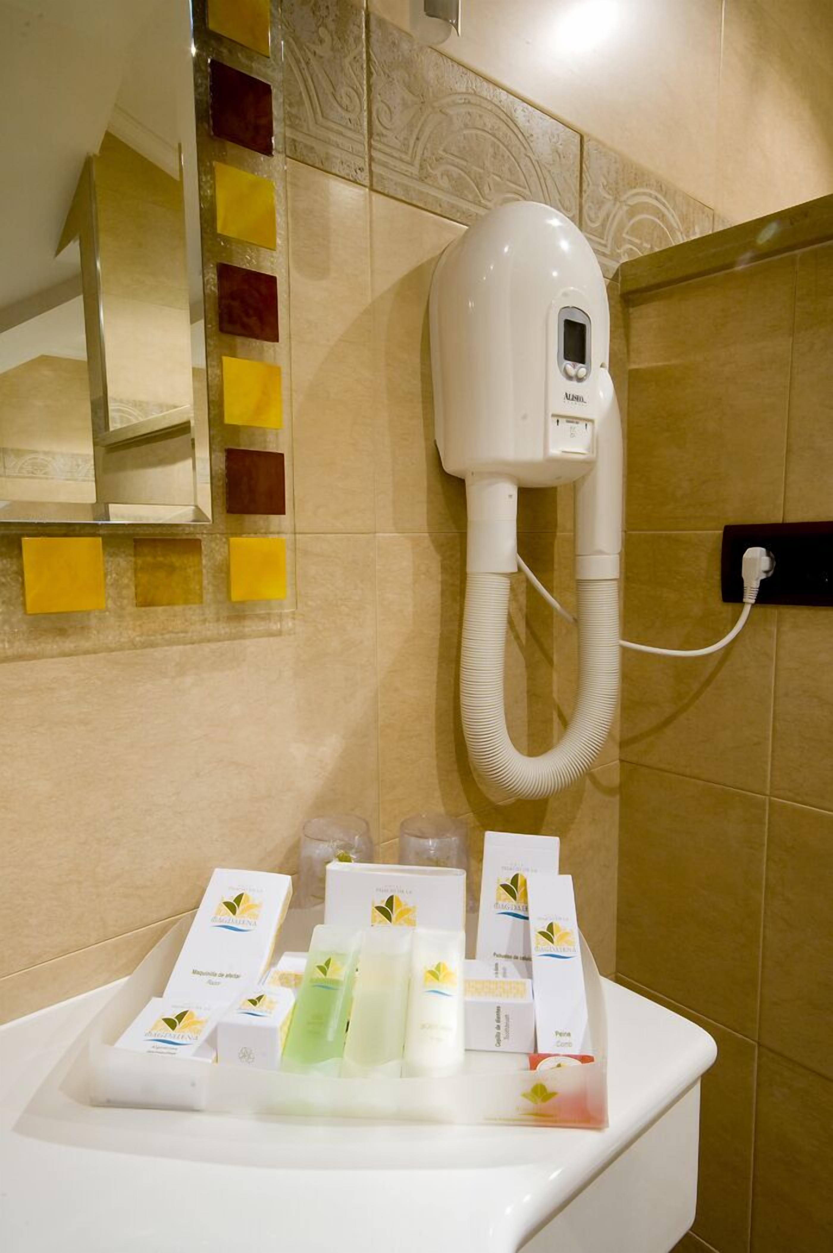 A bathroom with a hairdryer mounted on the wall, a sink with toiletries, and a mirror with decorative tiles.