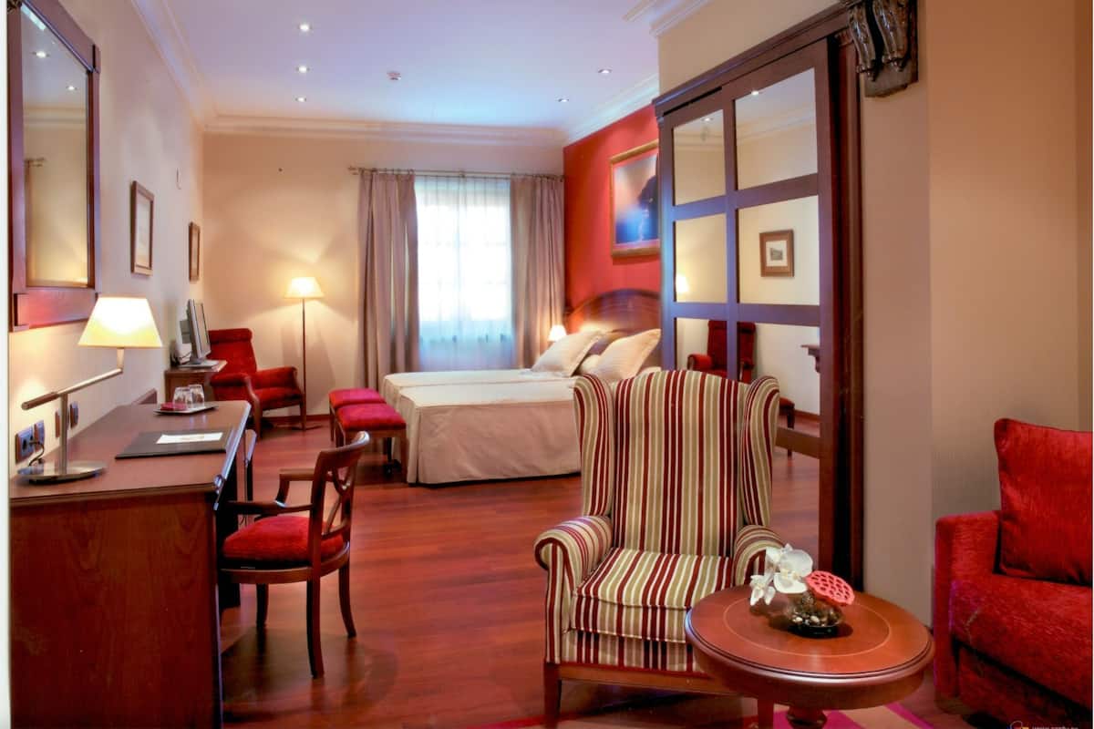 A hotel room with a bed, a desk, a chair, a striped armchair, and a small table with a decorative item.