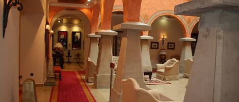 A hotel corridor with a red carpet, white armchairs, and stone pillars.