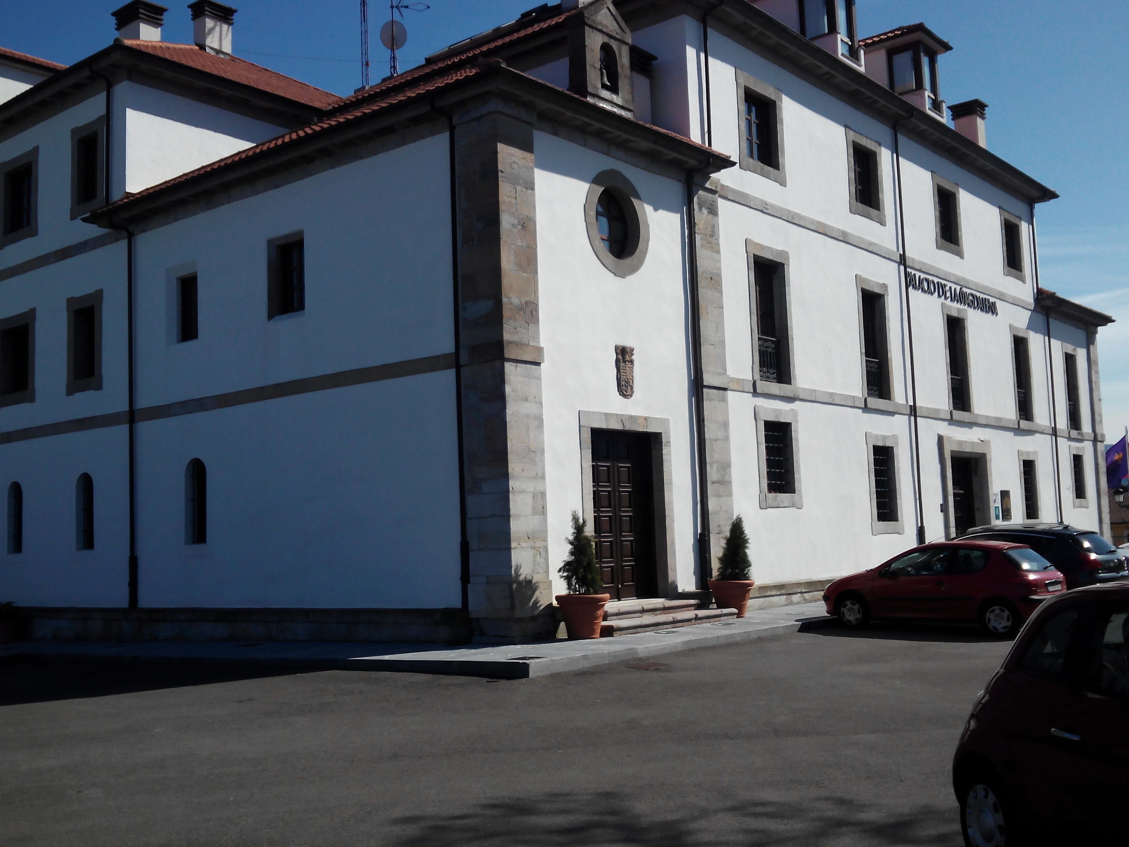 Un edificio bianco con tetto rosso, diverse finestre e un'entrata. Piante in vaso davanti e un'auto rossa parcheggiata nelle vicinanze.