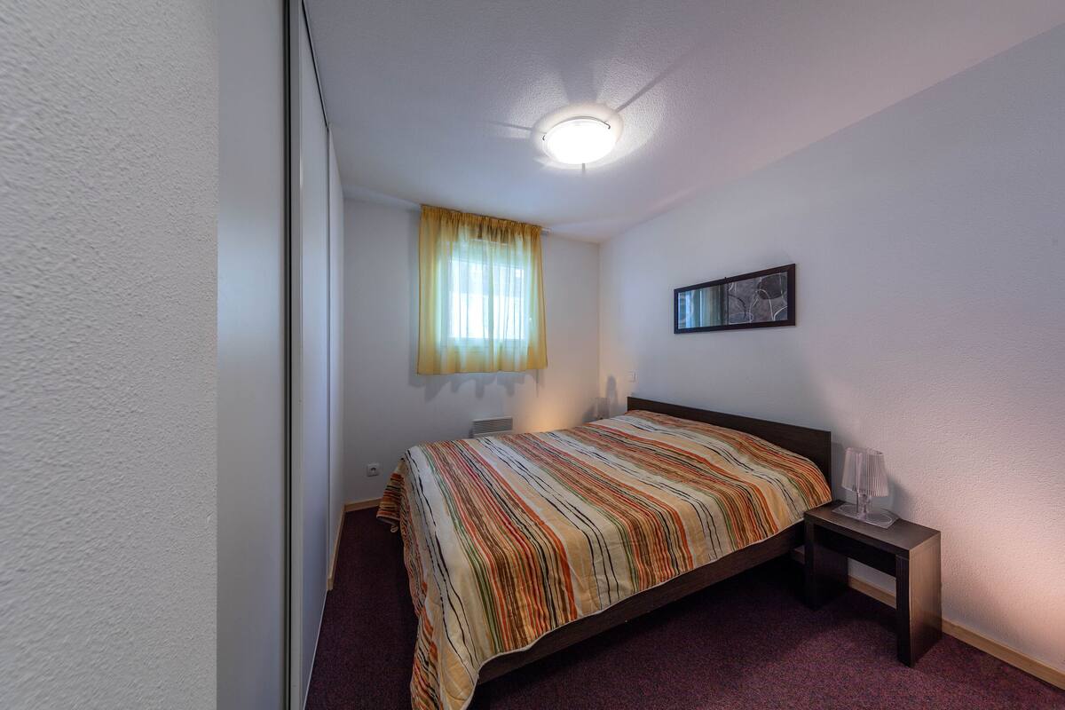 Quarto com uma cama de solteiro, mesa de cabeceira com abajur, janela com cortinas e luminária no teto.