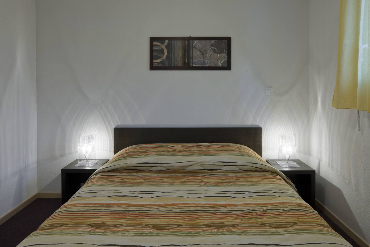 Quarto de hotel com cama, duas mesas de cabeceira, uma obra de arte na parede e uma janela com cortinas.