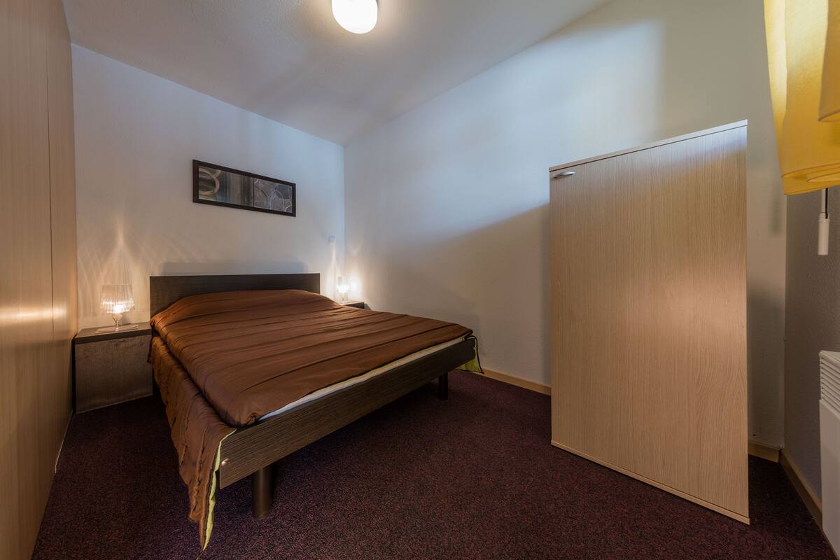 Quarto com uma cama de solteiro, mesa de cabeceira, luminária na parede, porta e radiador.