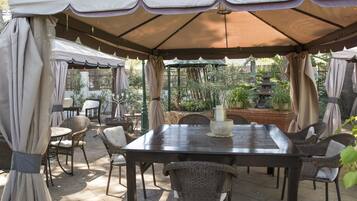 An outdoor patio with a table and chairs under a canopy, surrounded by greenery and a decorative fountain.