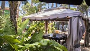 Un espacio al aire libre cubierto con una mesa y sillas, rodeado de grandes plantas tropicales.