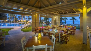 An outdoor dining area with tables and chairs, a pool in the background, and palm trees. - Aquamare Beach Hotel & Spa (Geroskipou)