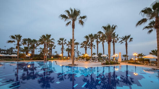 A swimming pool with tiled flooring, surrounded by palm trees and lounge chairs. - Aquamare Beach Hotel & Spa (Geroskipou)