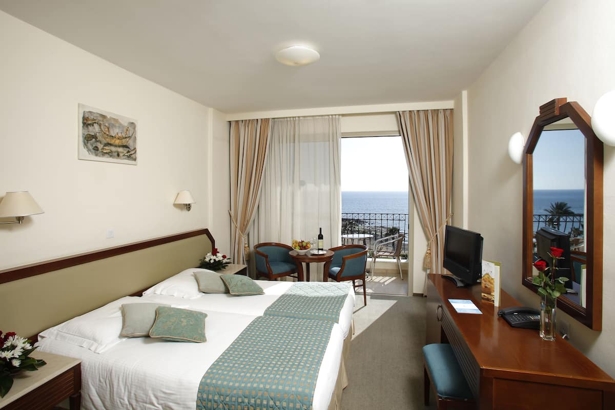 A hotel room with a bed, two chairs, a desk with a computer, a mirror, and a view of the sea.