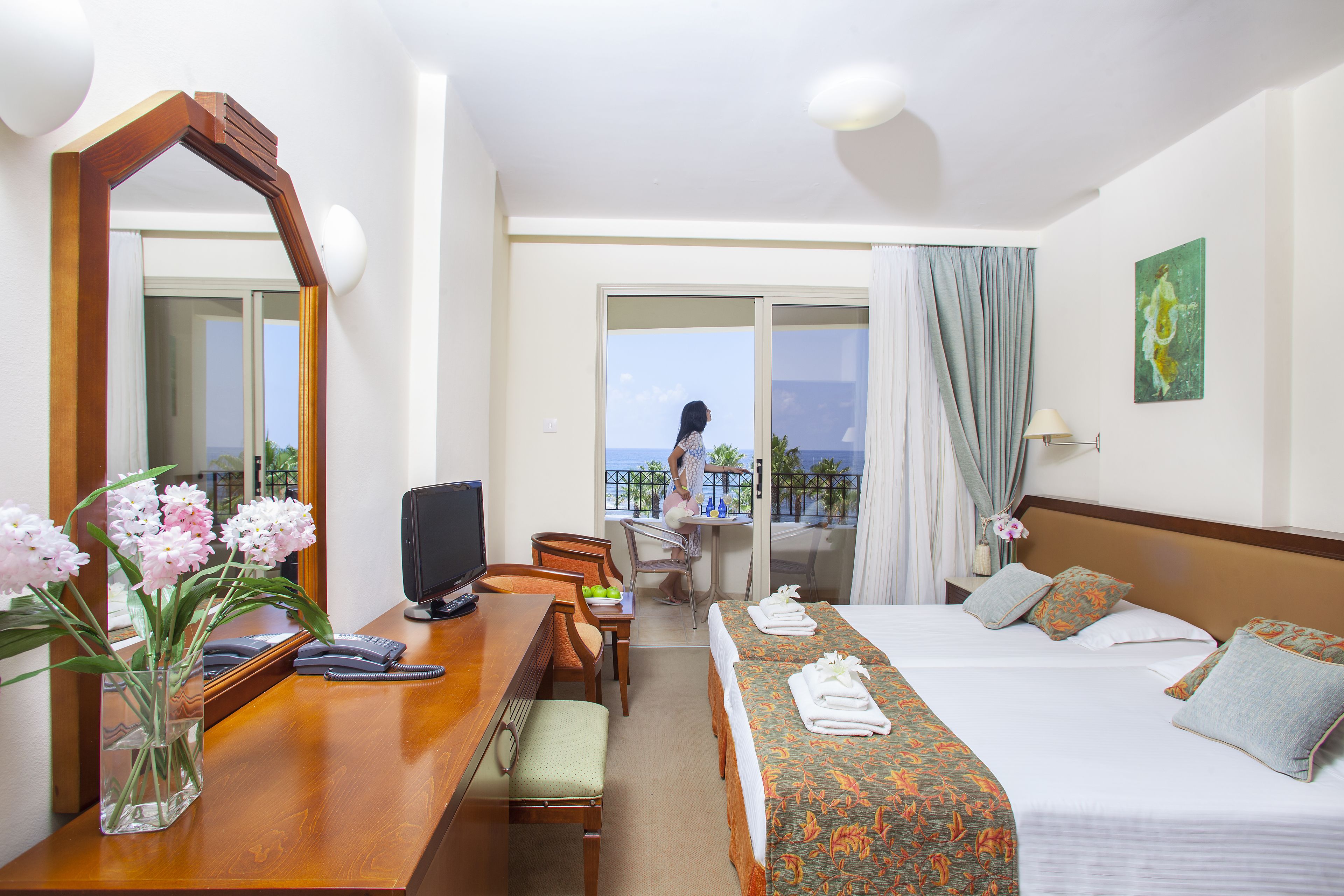 Habitación de hotel con una cama, un escritorio con computadora, una silla, un espejo y vistas al balcón.