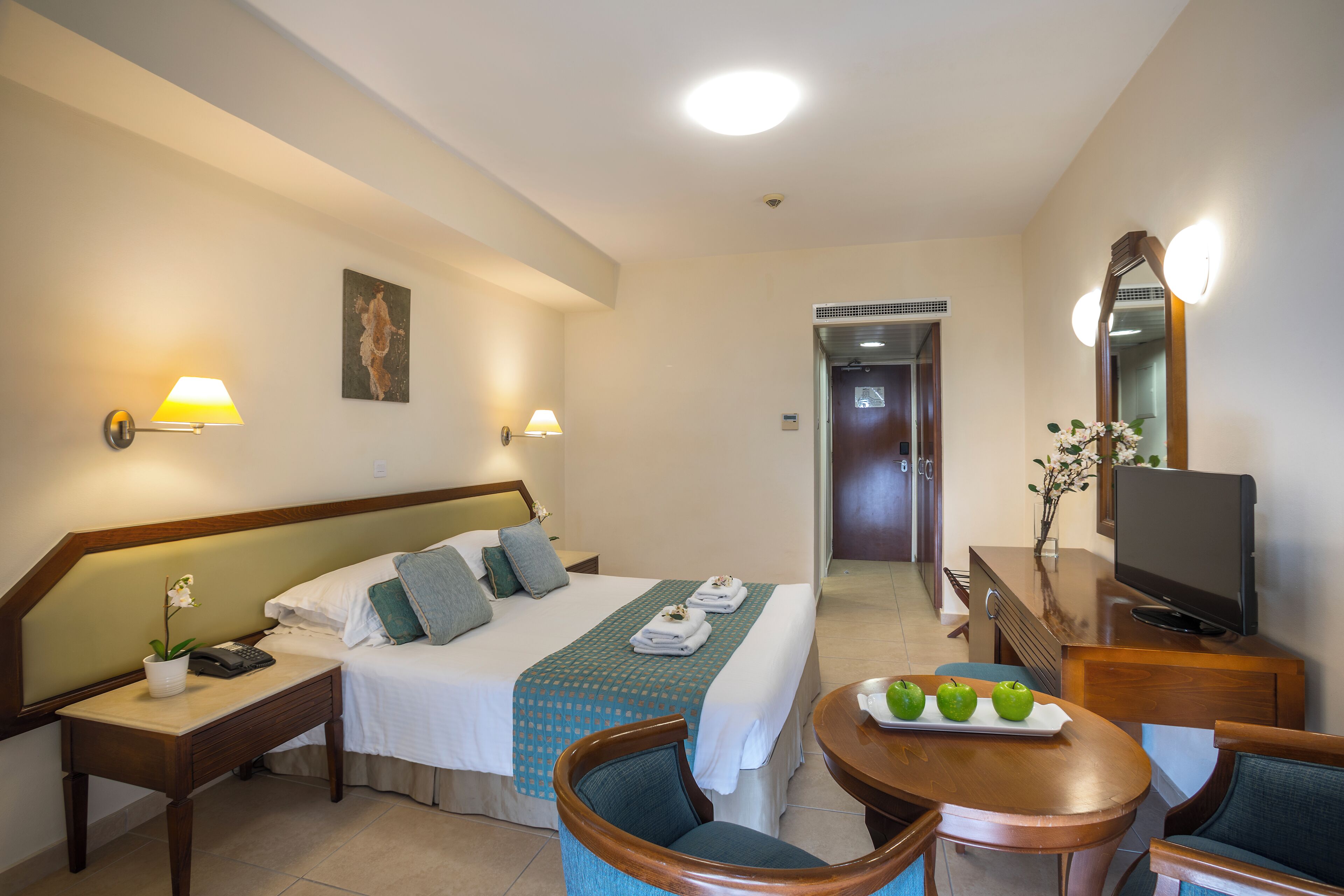 Ein Hotelzimmer mit Bett, Nachttischlampen, Schreibtisch mit Telefon, Fernseher und Essecke mit Tisch und Stühlen.