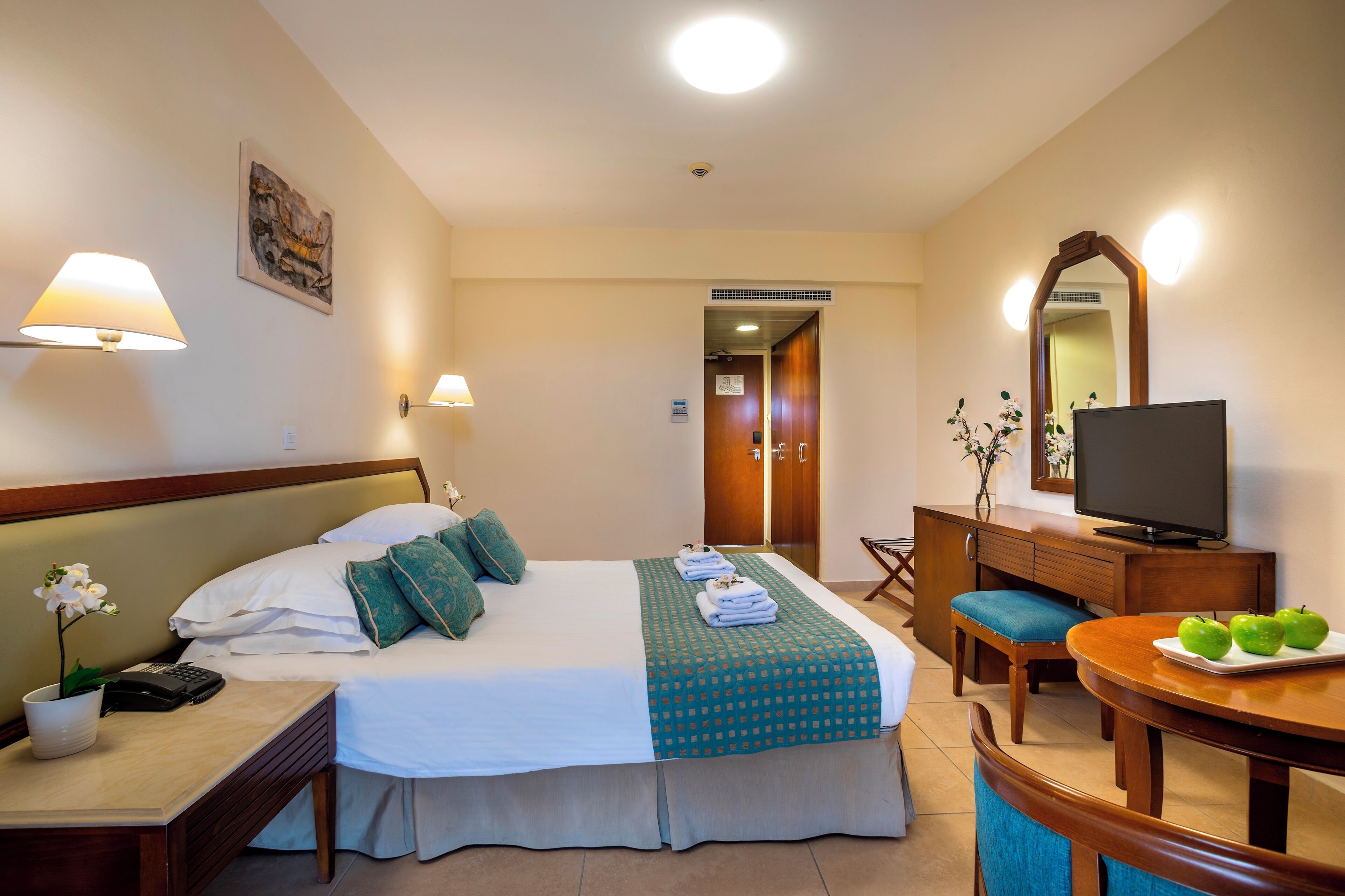 Ein Hotelzimmer mit einem Bett, Nachttischlampen, einem Schreibtisch mit Telefon, einem Fernseher und einem Esstisch mit Äpfeln.
