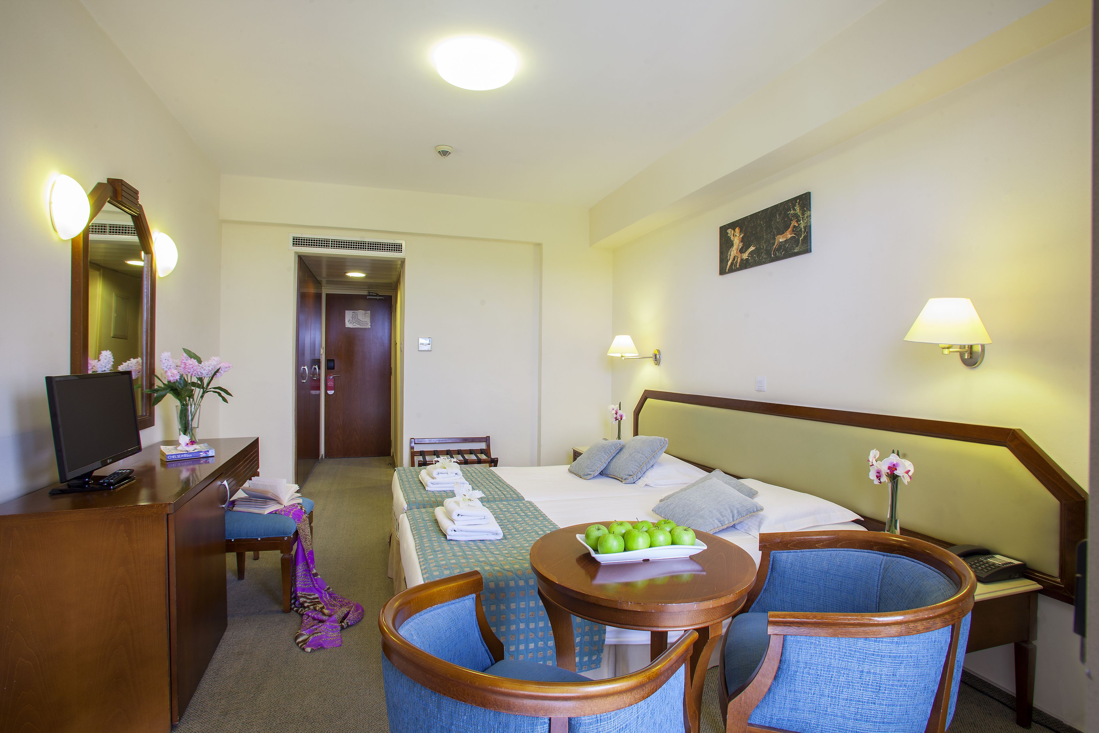 A hotel room with a bed, desk, chairs, a TV, and a small table with apples.