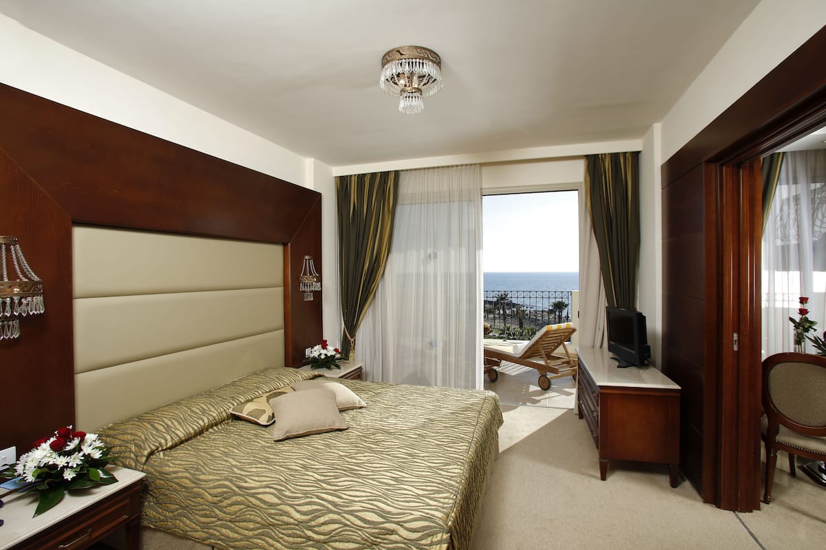A hotel room with a large bed, a desk with a television, a chair, and a view of the ocean.