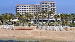 A beachfront hotel with multiple floors, palm trees, and beach umbrellas. - Aquamare Beach Hotel & Spa (Geroskipou)