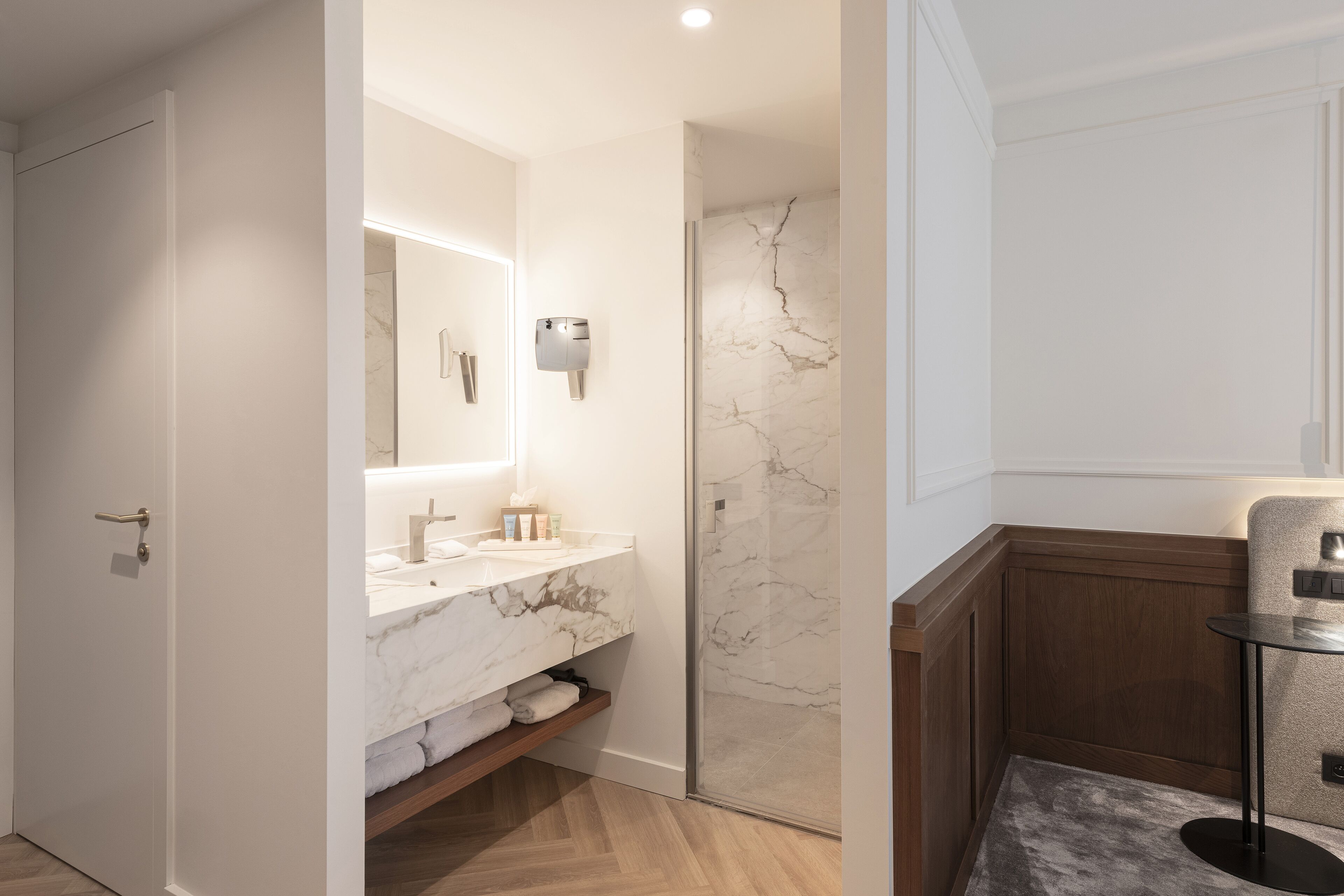 A modern bathroom with a marble vanity, a glass shower enclosure, and a small round table with a chair.
